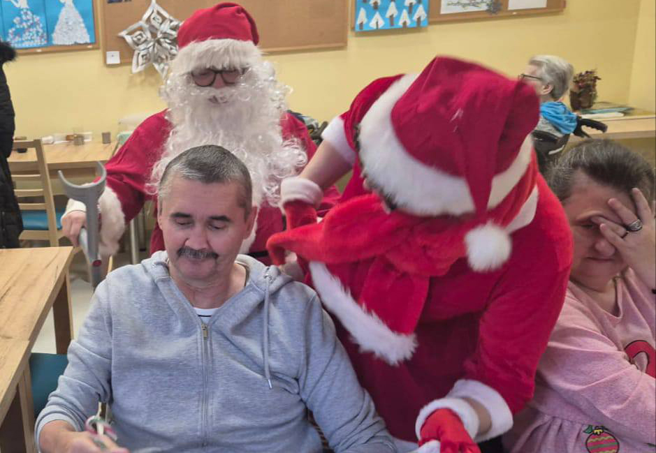 Mikołaj wręcza prezent mieszkańcowi Domu Pomocy Społecznej za nimi drugi Mikołaj