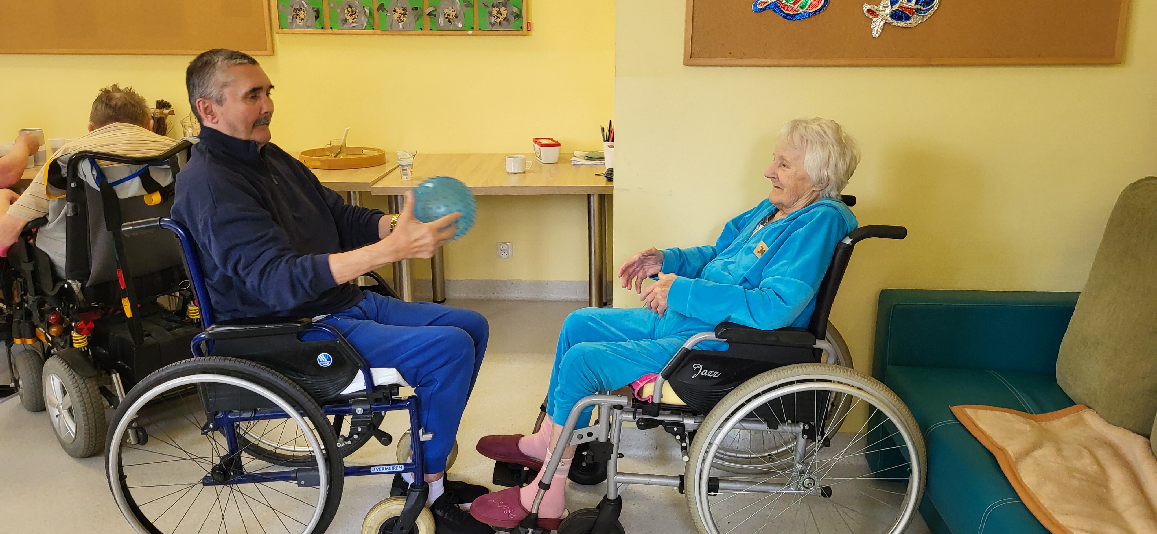 Dwoje seniorów na wózku ćwiczy podczas rehabilitacji rzucając do siebie piłka