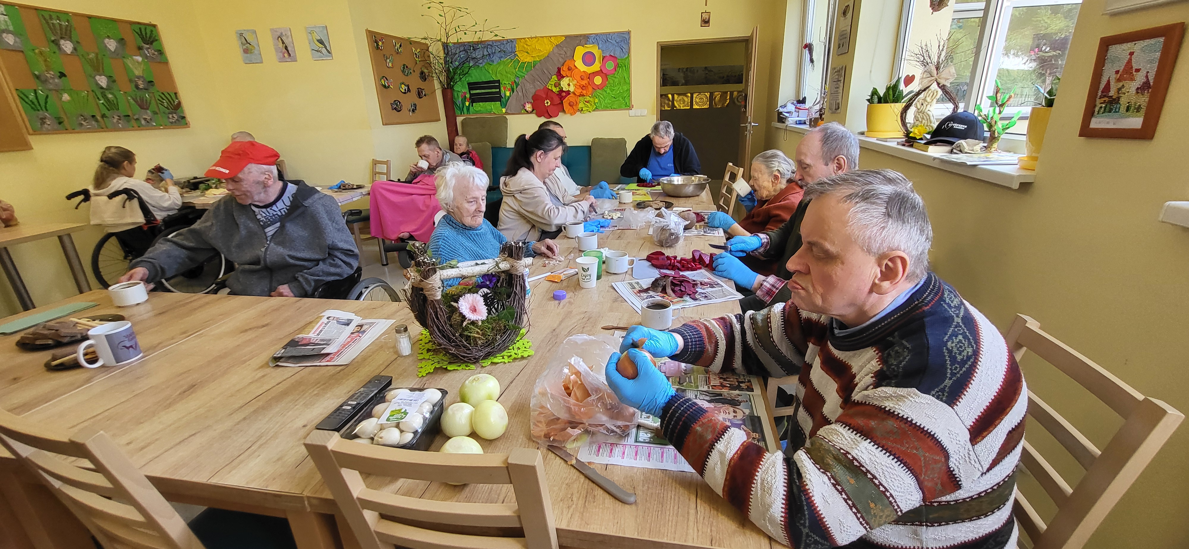 Mieszkańcy DPSu przy stole obierają warzywa na zajęcia kulinarne. Na pierwszym planie starszy Pan obierający cebule. 