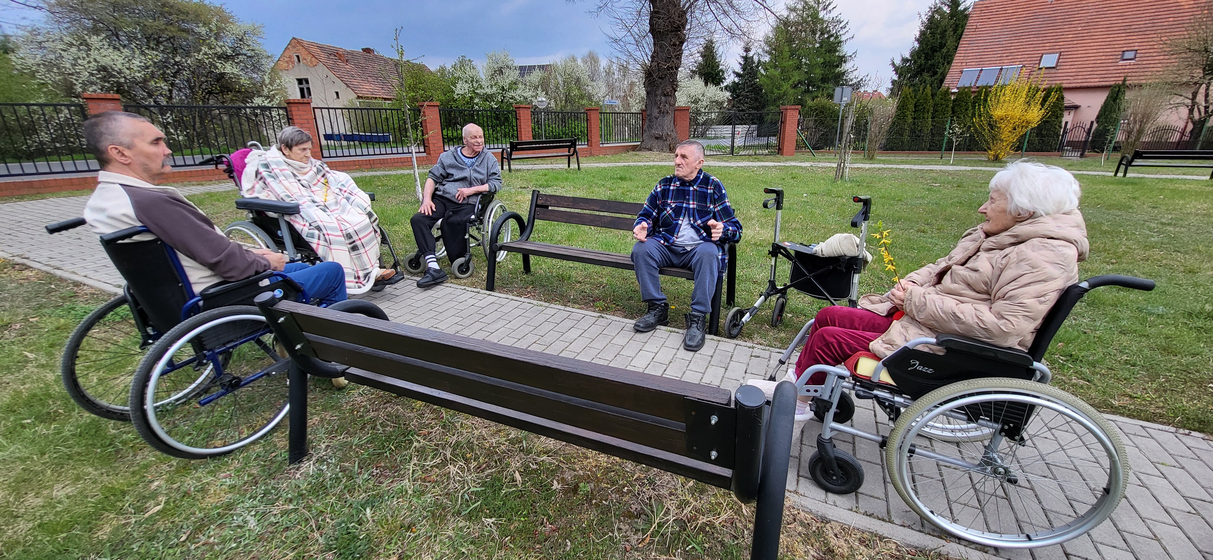 Mieszkańcy DPSu siedzą na ławkach i wózkach inwalidzkich łapią ciepłe promienie słońca